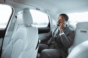 African American businessman in a grey suit is sitting in the backseat of a car, looking out the window while speaking on his phone. The car has a light-colored leather interior. photo