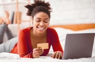 Online Banking. Black Lady Customer Using Credit Card And Laptop Computer Lying In Bed At Home On Weekend. Selective Focus photo