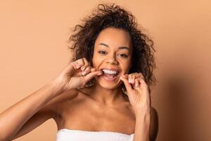 This image features African American woman with curly brown hair flossing her teeth in a studio setting. She is smiling and appears to be happy and confident. The background is a soft brown color. photo