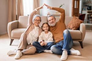 A smiling elderly couple and a young girl sit on the floor in a cozy living room, forming a house outline with their hands above them, symbolizing unity and family love. photo