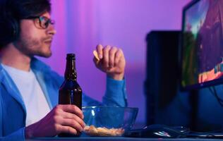 A young man in a blue shirt and glasses sits at a desk with a computer monitor in front of him. He is playing games while holding a bottle of beer in one hand and a bowl of chips photo