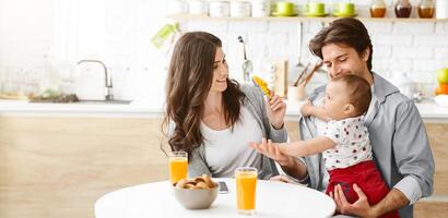 A happy family enjoys a meal together in their kitchen. The father holds their baby, who is reaching out to the mother who sits across the table, holding a yellow toy in her hand, copy space photo