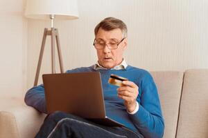 Online Shopping Concept. Elderly Gentleman Using Credit Card And Laptop Computer Buying Things On Internet Sitting On Sofa At Home photo