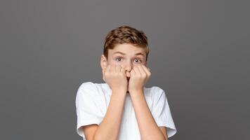 Teenager boy biting his nails in a pose reflecting anxiety and fear, isolated on a grey backdrop, capturing a common youngster behavior photo
