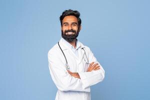 A confident Indian doctor is smiling warmly, dressed in a white lab coat with a stethoscope draped around his neck, standing against a plain blue background. photo