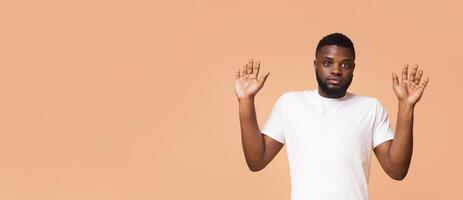 A young Black man in a white T-shirt with short hair and a beard stands in front of a light background. He is looking at the camera with a serious expression and has both hands raised in front of him photo