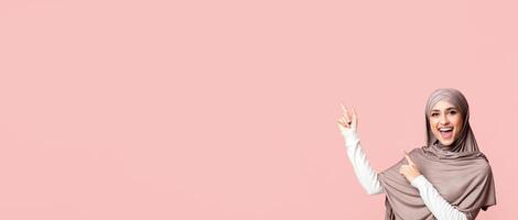Great offer. Cheerful arabic girl in hijab pointing at copy space on pink background with two hands, showing or advising something, panorama with empty space photo