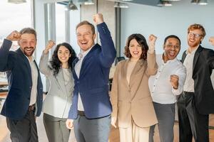 A group of diverse business professionals stand in a modern office, all smiling and raising their fists in celebration. They appear to be celebrating a successful project or achievement. photo