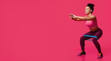 Young sporty smiling black woman doing stretching work out with elastic bands, squating over red studio background photo