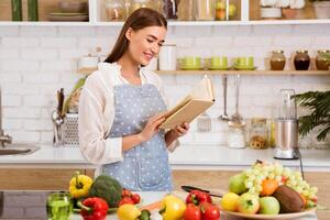 A woman stands in her kitchen wearing an apron and reading a cookbook. She is preparing to cook a meal, as evidenced by the fresh produce on the counter. photo