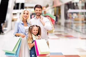 A young family, consisting of a father, mother, and daughter, are standing together in a modern mall, all smiling and holding colorful shopping bags. photo