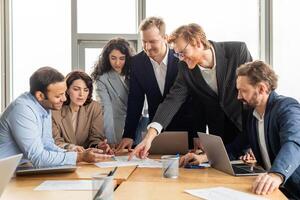A group of business professionals are gathered around a table in a modern office, collaborating on a project. They are reviewing documents and using laptops. photo