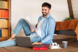 A cheerful man in casual attire sits on the floor with his laptop open and paperwork in hand, sitting area features a coffee cup, headphones, and books, suggesting a relaxing work-from-home setup photo