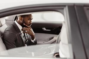 Black businessman, dressed in a suit and tie, sits in the backseat of a car, working on a laptop. He has a thoughtful expression on his face as he focuses on his work photo