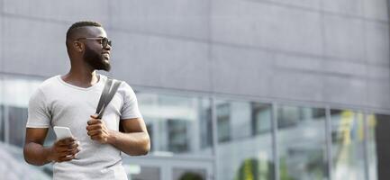A man in sunglasses and a white t-shirt walks in a city with a phone in his hand, looking to his right. photo