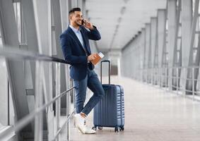 Mobile Call. Smiling Young Middle Eastern Businessman Talking On Cellphone In Airport While Waiting With Suitcase For Flight At Terminal, Happy Arab Man Having Pleasant Phone Conversation photo