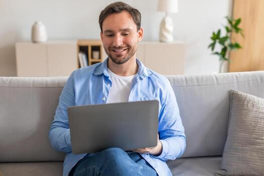A man with a smile on his face is comfortably seated on a couch, engrossed in using a laptop computer. He appears relaxed and focused on his work, with the glow of the screen illuminating his face. photo