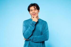Asian young guy wearing a casual blue sweater places one hand on his chin, striking a thoughtful pose. He stands against a light blue background, creating a calming and serene atmosphere. photo