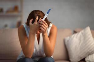 Unwanted pregnancy. Unrecognizable woman holding test covering face in despair sitting on sofa at home. Selective focus photo