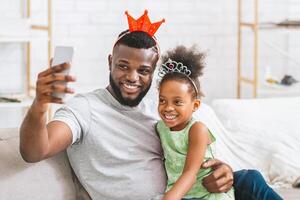 A happy black father and daughter, both wearing crowns, sit on the couch together and take a selfie with a smartphone. The father is smiling and holding the phone out in front of him. photo