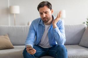 A man in casual blue attire appears perplexed and somewhat distressed as he holds his smartphone in one hand, gesturing with the other as if struggling to understand a message or situation photo