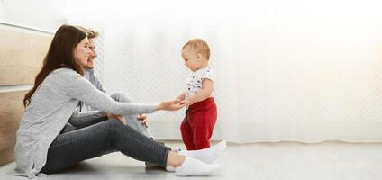 A young family of three are spending time together at home. The parents are sitting on the floor, smiling at their baby who is taking their first steps, copy space photo