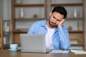A young man sits at a wooden table, looking troubled as he leans his cheek on his hand with a closed laptop in front of him. His expression suggests dealing with a challenging problem photo