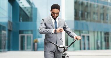 A professional black man on a bicycle is looking at his watch in an urban area. The scene is modern and energetic, showcasing the blend of professional responsibilities and efficient commuting. photo
