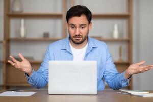 A man in a casual blue shirt sits at a desk, showing a bewildered expression with arms raised in uncertainty as he looks at his laptop screen, likely dealing with a perplexing work situation photo
