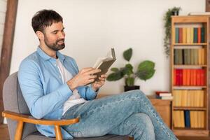 A man is seated in a chair, engrossed in reading a book. The mans attention is focused on the pages of the book, his posture relaxed as he flips through the pages. photo