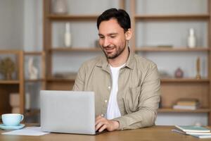 A man is seated at a table, focused on his laptop screen. Beside him, there is a steaming cup of coffee. The man appears engrossed in his work, typing on the laptop keyboard while sipping his coffee. photo