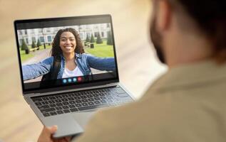 African American woman is on a call with someone on a laptop computer. She is smiling and waving, and appears to be in a park setting. She is likely calling a friend photo