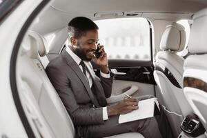 African American businessman in a suit sits in the backseat of a luxury car and makes notes on a notepad while speaking on his phone. He looks focused and professional. photo