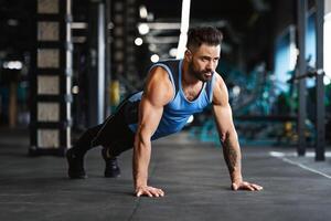 A man with a beard is doing push-ups in a gym. He is wearing a blue tank top and black pants. photo