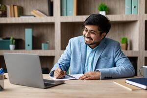 A smiling Indian man in a blue suit is sitting at a desk, taking notes in a notebook while looking at his laptop. The man is focused on his work and appears happy. photo