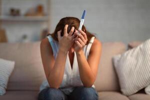 Unrecognizable Depressed Girl Holding Pregnancy Test Covering Face With Hands Sitting On Couch Indoor. Selective Focus photo