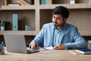Indian man in a blue blazer and glasses sits at a wooden desk, writing in a notebook with a blue pen. His laptop is open in front of him, and there are a few other notebooks and a mug on the desk. photo
