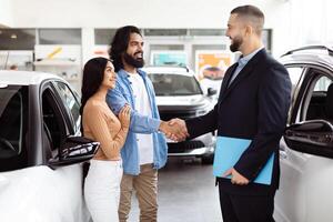 A young Indian couple is finalizing a deal with a car salesman inside a vehicle showroom. They seem pleased and are shaking hands with the agent, who is dressed in a business suit photo