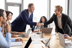 A group of business professionals are gathered around a conference table in a modern office setting. Two of the professionals are standing and shaking hands, while the others sit and watch photo