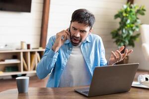A man is seated at his work desk at home, expressing frustration while speaking on his mobile phone. He appears agitated, gesturing with his free hand photo