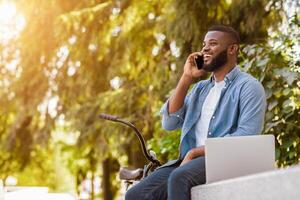 Cell Communication. Portrait Of Happy Handsome Afro Man Talking On Cellphone While Sitting With Laptop And Bicycle In Park photo
