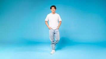 Asian guy in a white t-shirt is standing upright in front of a plain blue background. His posture is relaxed, with arms by his side and feet grounded on the floor. photo