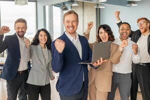 A diverse group of business professionals stand together in a modern office, celebrating a successful project. Team is enthusiastic, with their arms raised in the air and bright smiles on their faces photo