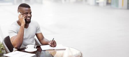 A man sits at a cafe table, talking on his phone and writing in a notebook, smiling while on a call. photo