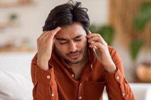 Arab man with dark hair and a beard is on a phone call inside. He has a worried expression and is holding his forehead with his left hand. The man is wearing a rust colored button up shirt. photo
