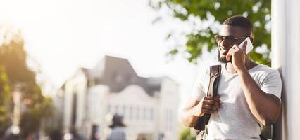 A man wearing sunglasses and a backpack is smiling while talking on a cell phone on a sunny day in the city. photo