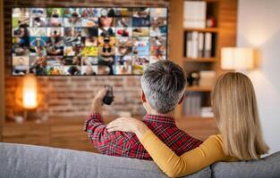 A couple is relaxing on a couch in their living room. The man is holding a remote control and is facing a large TV screen that is displaying a grid of smaller images. photo