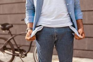 Poverty and absence of money. Unrecognizable afro man demonstrating empty turned out pockets outdoors, standing next to his bike, cropped image photo