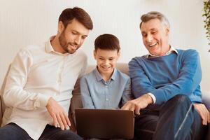 Male Family. Father And Grandfather Helping Boy Do Homework On Laptop Computer Sitting On Sofa At Home photo