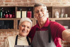 An elderly couple, wearing aprons, is filming a cooking show in their home kitchen. The man is in the foreground, gesturing towards the camera while smiling, taking selfie photo